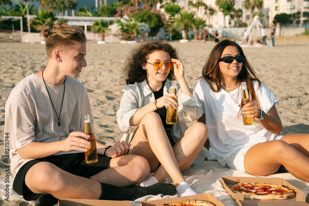 Young happy friends doing picnic seaside close-up portrait. Group of people sitting on the beach, eating pizza and drinking beer together. Summer spending time outdoor concept.