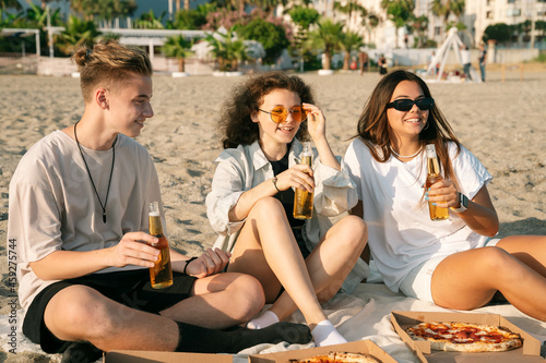 Young happy friends doing picnic seaside close-up portrait. Group of people sitting on the beach, eating pizza and drinking beer together. Summer spending time outdoor concept. © valeriyakozoriz
