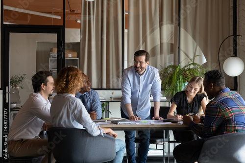 Happy male business team leader talking to employees at corporate meeting, discussing work project, sharing jokes, laughing. Positive mentor training diverse group of interns, having fun