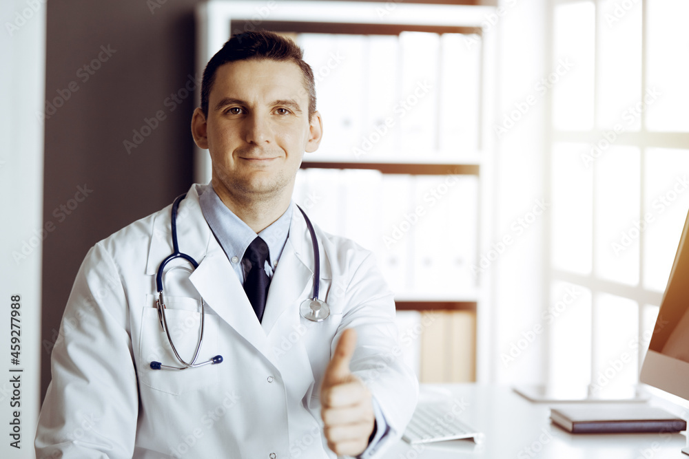 Friendly male doctor sitting and showing OK sign with thumbs up at his working place in sunny clinic. Perfect medical service in hospital