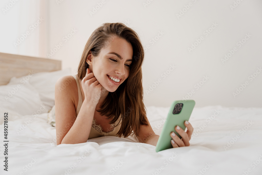 Fun young woman in casual casual clothes lying on her stomach hold in hand use watch mobile cell phone get video call wave hand rest relax spend time in bedroom lounge home in own room house good day. Fun young woman in casual casual clothes lying on her stomach hold in hand use watch mobile cell phone get video call wave hand rest relax spend time in bedroom lounge home in own room house good day.
