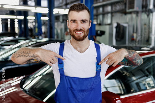 Strong smiling young male professional technician car mechanic man in denim blue overalls white t-shirt point on himself work in light modern vehicle repair shop workshop indoors Tattoo traslate fun. © ViDi Studio