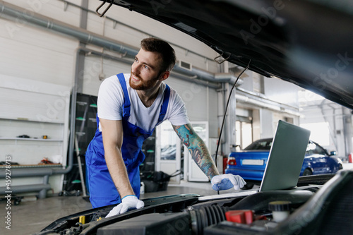 Young smiling professional technician car mechanic man in blue overalls t-shirt use laptop pc computer make diagnostics check fix problem with raised hood work in vehicle repair shop workshop indoor