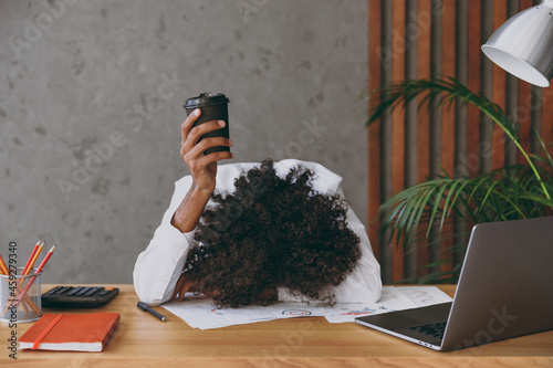 Young sad african american employee business man wearng classic shirt put head down on table sleeping hold takeaway delivery craft paper cup coffee sit work at white office desk with pc laptop inside.