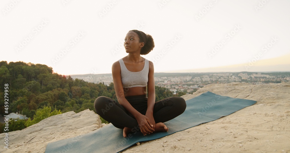 Young multiracial woman sitting in yoga posture and meditating at the mountains. Girl performing aerobics exercise and morning meditation at the nature. Physical and spiritual practice concept
