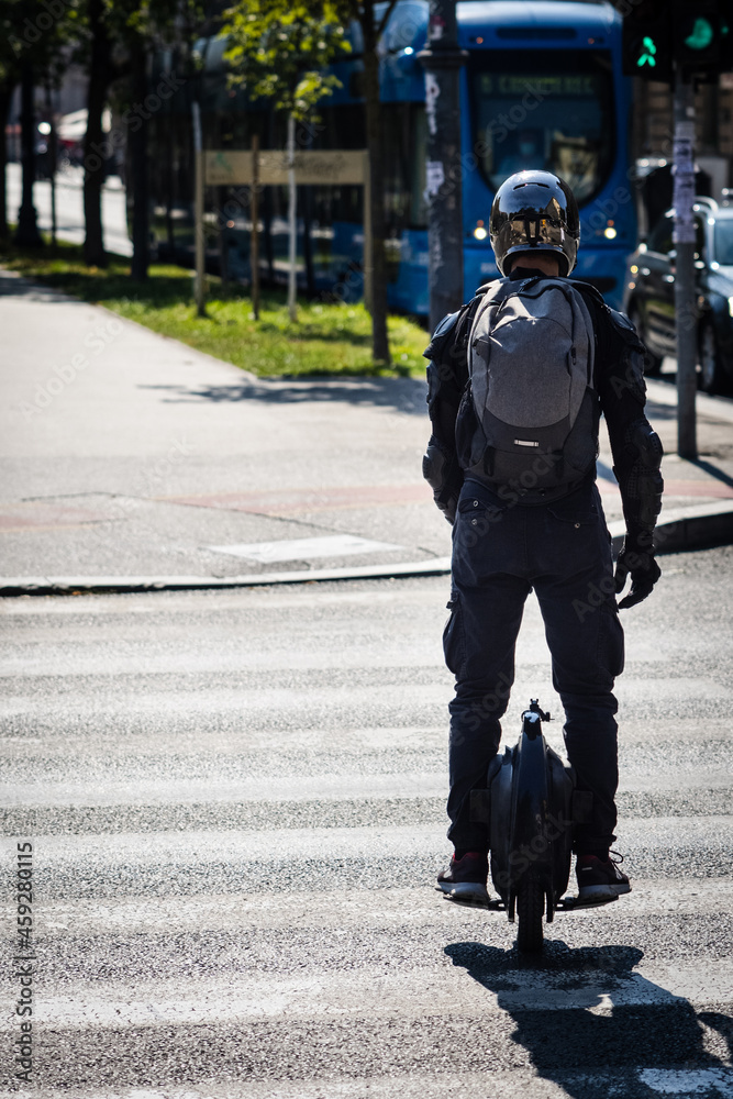 Obraz premium Fully equipped man driving on electric monowheel in urban city area, crossing street on a crosswalk, rear view