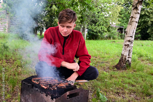 young man roasts meat, barbecue on coals on grill in nature. He's outside. Barbecue is located on green grass. There are trees in background. Concept of spending free time. Copy space