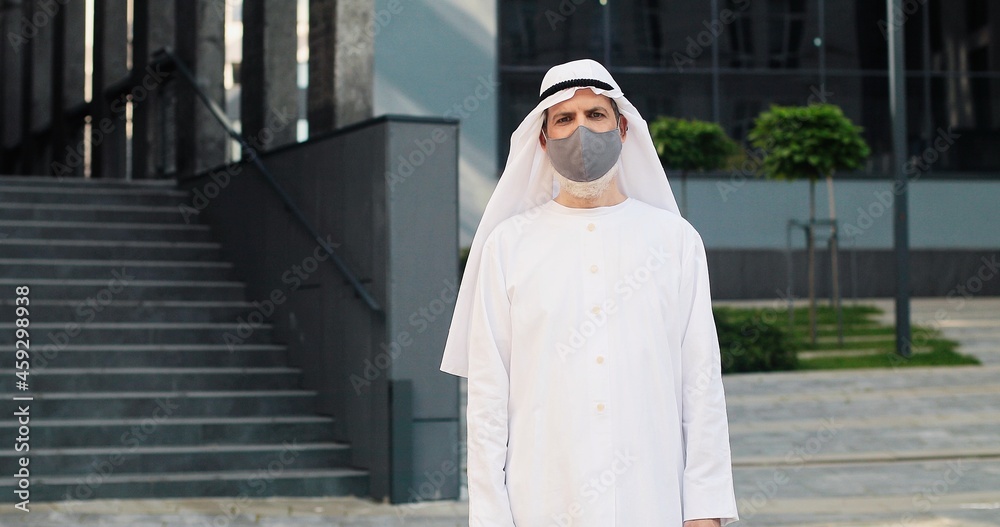 Pandemic period. Waist up portrait view of the senior sheikh wearing protective mask standing at the street with office center at the background. Quarantine concept