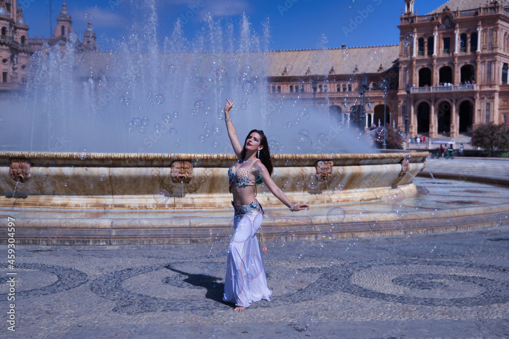 Obraz premium Middle-aged Hispanic woman, wearing a beautifully colored dress with rhinestones, for belly dancing, dancing surrounded by soap bubbles in front of a water fountain. Belly dance concept.