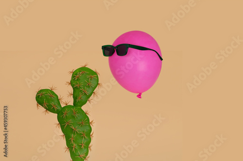 Cactus plant with above it floating a pink balloon in sunglasses on a beige background.