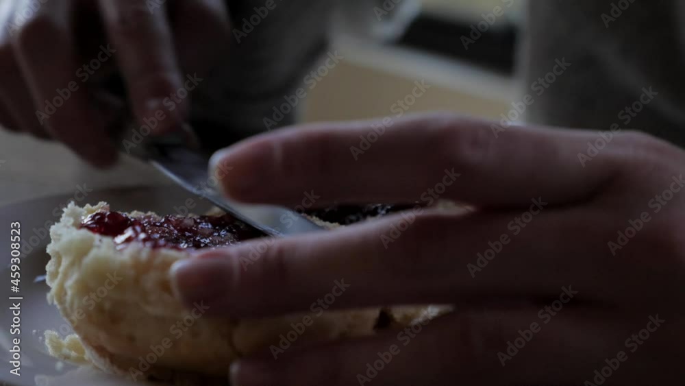 Female hand spreads strawberry raspberry jam from one freshly baked traditional scone to another and uses knife for afternoon tea snack. Female hand spreads strawberry raspberry jam from one freshly baked traditional scone to another and uses knife for afternoon tea snack.