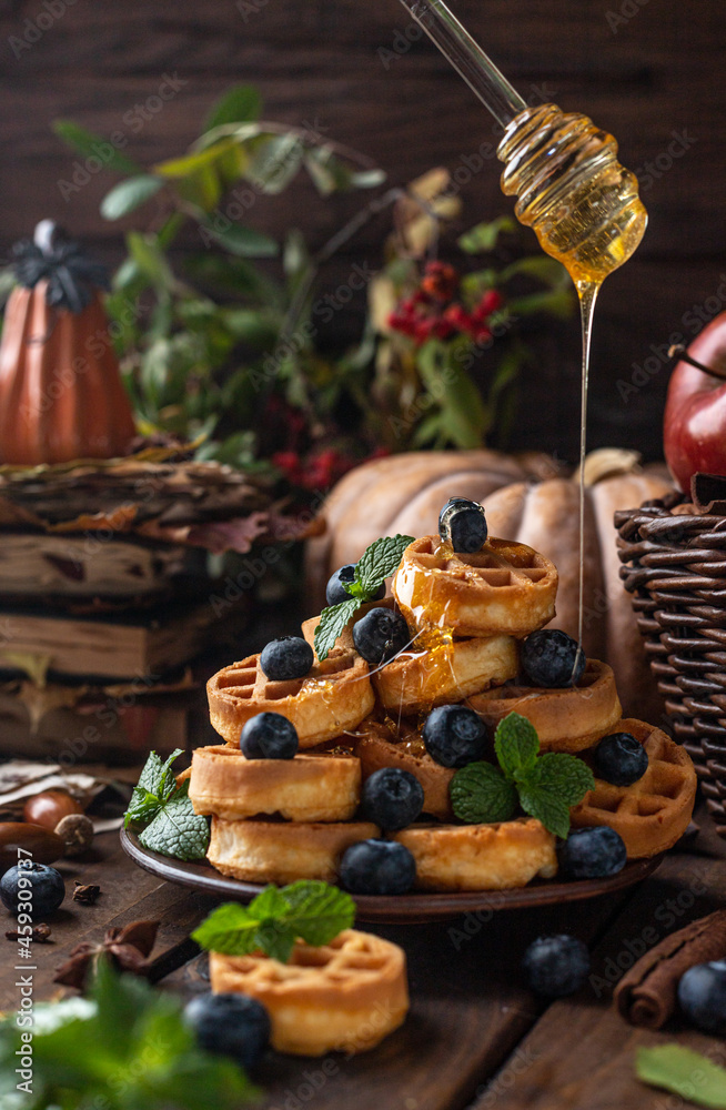 many waffles are stacked on a plate, decorated with mint leaves and blueberries. On top of them, honey pours from glass spoons