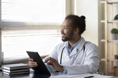 Happy millennial Afro American doctor looking at window away, smiling, holding tablet, thinking over distance online consultation. Practitioner using gadget for reading electronic medical records