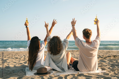 Young happy friends doing picnic and cheers with bottles seaside. Group of people drinking beer together on the beach.