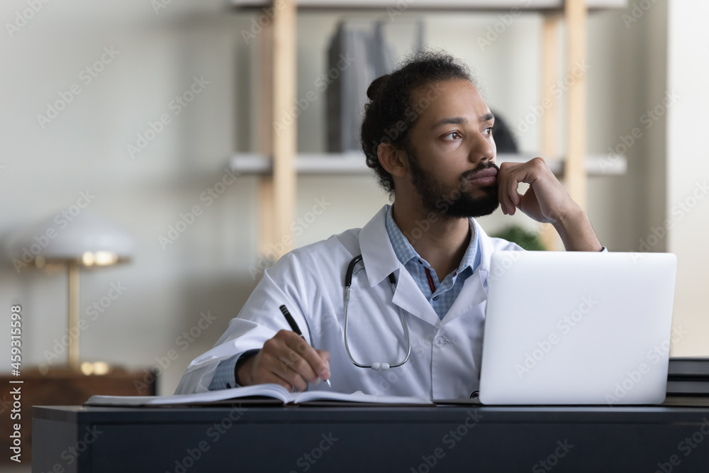 Thoughtful serious male mixed race doctor looking at window in office, thinking of patients, future vision, medical career growth, promotion, working at table with laptop, writing records. Thoughtful serious male mixed race doctor looking at window in office, thinking of patients, future vision, medical career growth, promotion, working at table with laptop, writing records.