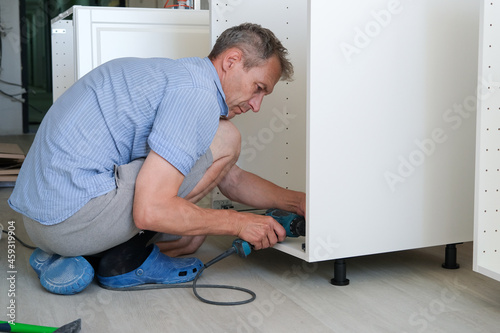 The man collects white kitchen furniture on his own. A man sits on the floor and screws it down with a screwdriver. Horizontal photo. Blue shirt and gray shorts.