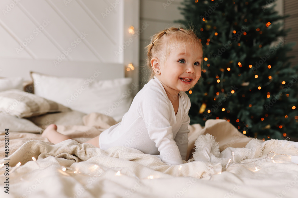 Cute funny baby little girl in pajamas smiling playing with a garland lying resting on bed linen on the bed by the Christmas tree in a decorated bedroom in a cozy house in the winter holidays indoors Cute funny baby little girl in pajamas smiling playing with a garland lying resting on bed linen on the bed by the Christmas tree in a decorated bedroom in a cozy house in the winter holidays indoors