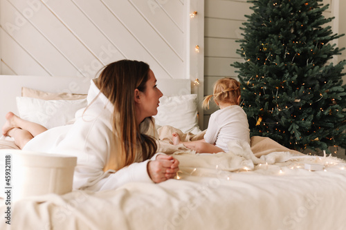 A young mother and a sad, touchy child are sitting on a bed in a bedroom with a Christmas tree in a cozy house waiting for the Christmas holidays and New Year. Family education. Selective focus