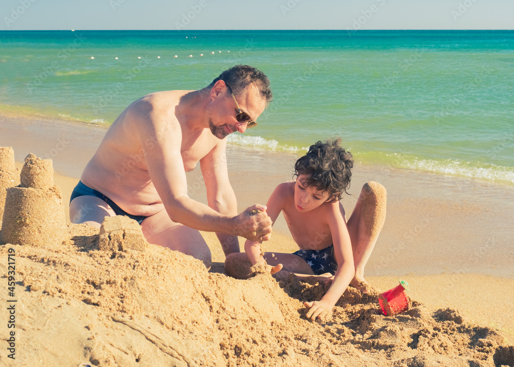 Bearded dad and little son are playing on the beach in the sand. Sea vacation on the shore on a sunny day. Bearded dad and little son are playing on the beach in the sand. Sea vacation on the shore on a sunny day.