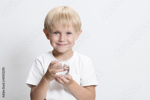 Cute Caucasian child, blond boy with glass of water. White studio background. Concept of clean water that can be drunk by children, world problem of water resources and drinking water. Copy space