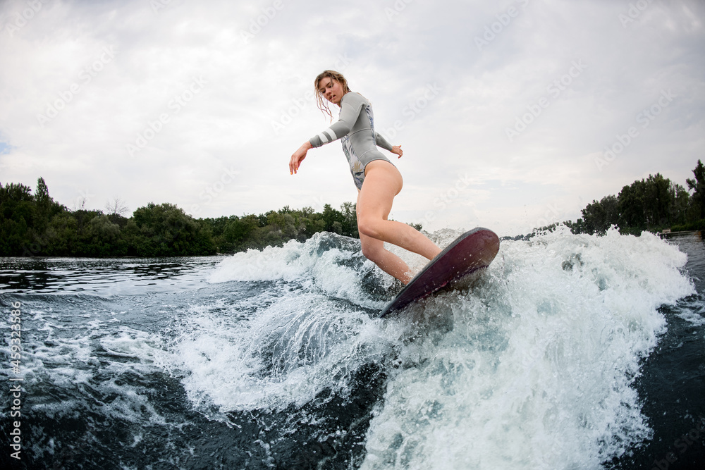 Flexible woman in grey swimsuit skilfully riding on wave on wakesurf board.