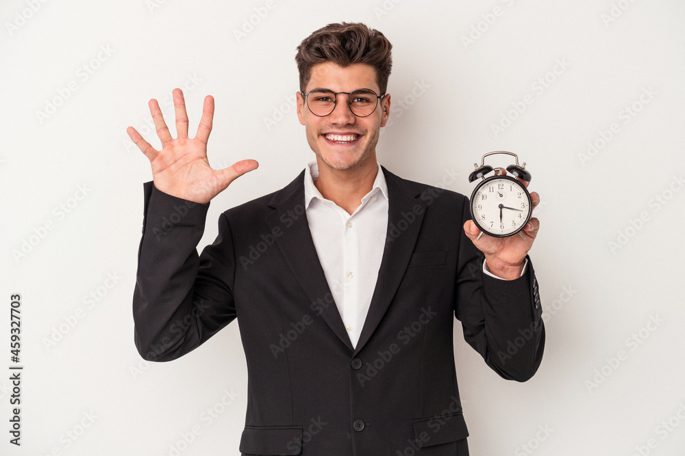 Young business caucasian man holding alarm clock isolated on white background smiling cheerful showing number five with fingers. Young business caucasian man holding alarm clock isolated on white background smiling cheerful showing number five with fingers.