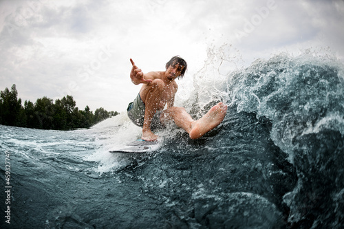 Active wet man balancing on wake surf board stretching his leg and showing hand gesture