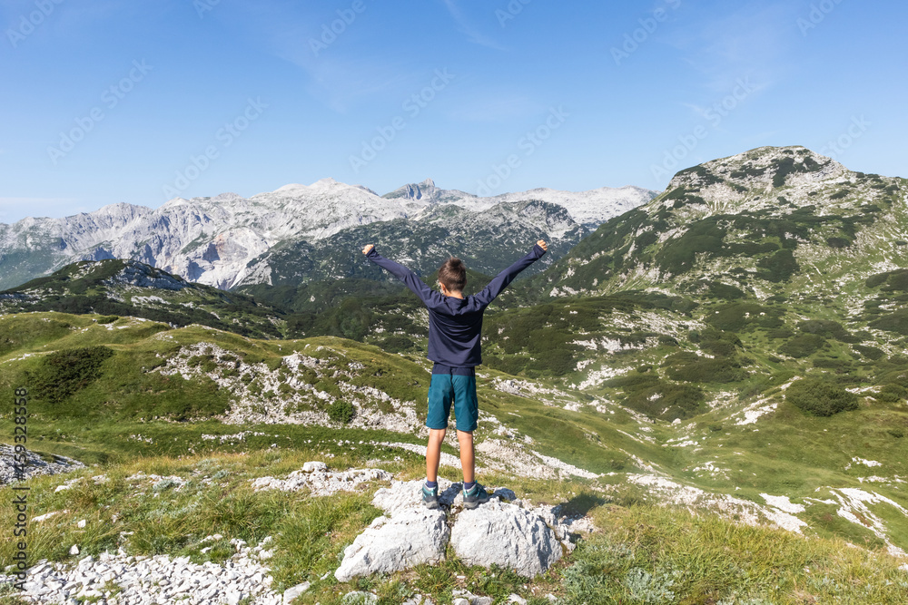 School-aged boy standing at the top of the hill, and looking at mountain tops, raising arms with fist pump gesture, celebrating mountaineering success