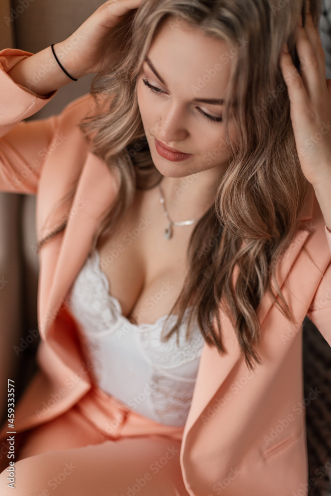 Obraz premium Beautiful young sexy stylish woman in a fashionable suit and blazer with white lace lingerie sits and rests in a chair