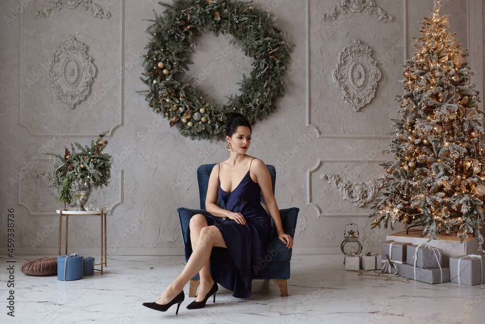 Adorable woman in a stylish dark-blue dress in front of Christmas tree. Lovable female model in trendy dress posing in the interior decorated for New year holidays - obrazy, fototapety, plakaty 