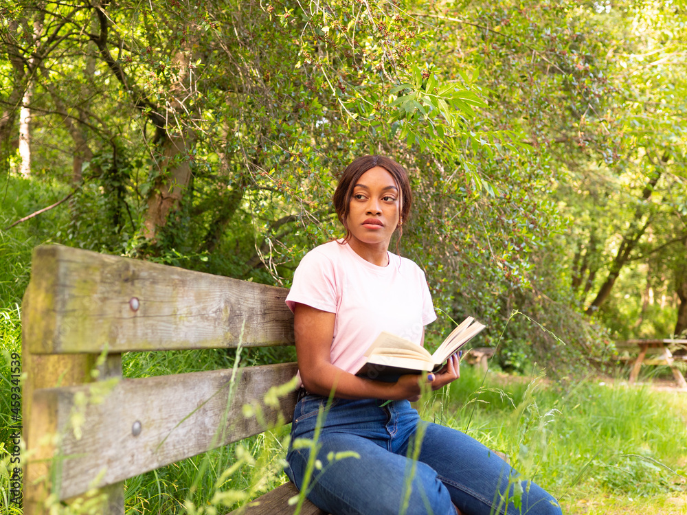 Young black woman sitting on a park bench reading a book.