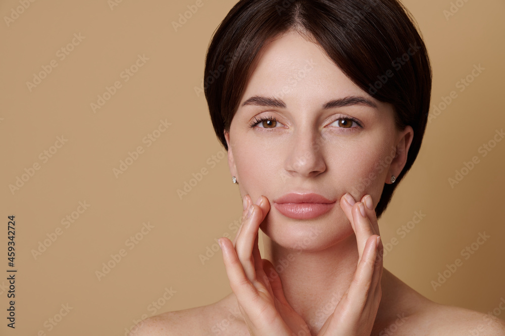 Beauty portrait of half naked European young gorgeous woman massaging her face, looking confidently at the camera isolated on beige background with copy space. Skin care, cosmetology and spa concept Beauty portrait of half naked European young gorgeous woman massaging her face, looking confidently at the camera isolated on beige background with copy space. Skin care, cosmetology and spa concept