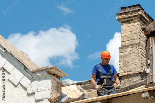 Repair of a wooden roof outdoors against the background of blue sky and clouds. A fashionable carpenter in sunglasses and special clothes with a hammer in his hand installs beams and boards.