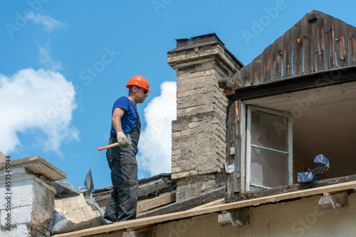 Repair of a wooden roof outdoors against the background of blue sky and clouds. A fashionable carpenter in sunglasses and special clothes with a hammer in his hand installs beams and boards.