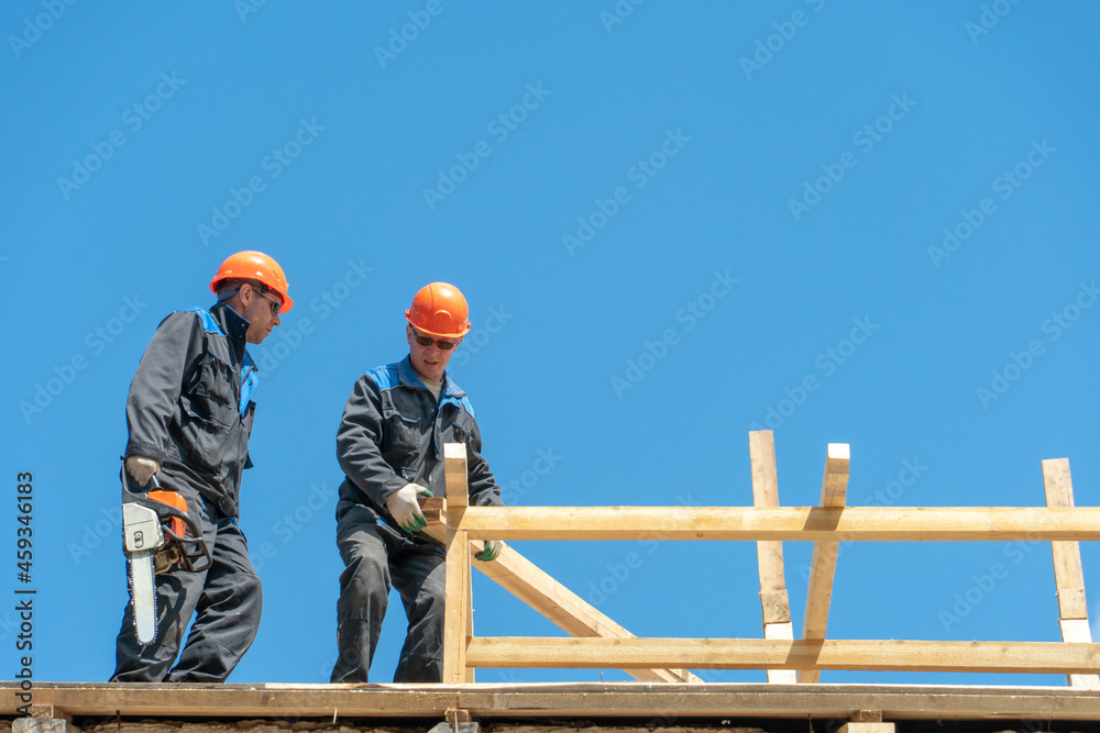 Repair of a wooden roof outdoors on a summer day against the background of blue sky and clouds. A carpenter in special clothes and with a tool installs beams and wood boards. Repair of a wooden roof outdoors on a summer day against the background of blue sky and clouds. A carpenter in special clothes and with a tool installs beams and wood boards.