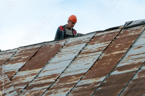 Repair of an old roof on the street. A view of an old tin roof made of rotten and rusty metal plates. The sad face of a worker dissatisfied with poor working conditions. A carpenter in a helmet.
