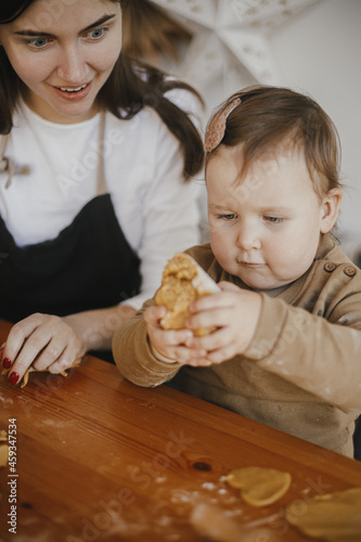 Adorable baby girl helping mother kneading dough for gingerbread cookies on wooden table in modern scandinavian room. Cute toddler daughter helps making christmas cookies. Authentic moment