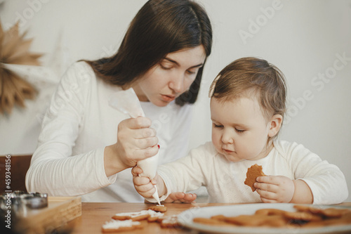Cute little daughter and mother together decorating christmas gingerbread cookies with icing on wooden table in modern room. Mommy daughter authentic lovely moments. Holiday preparations