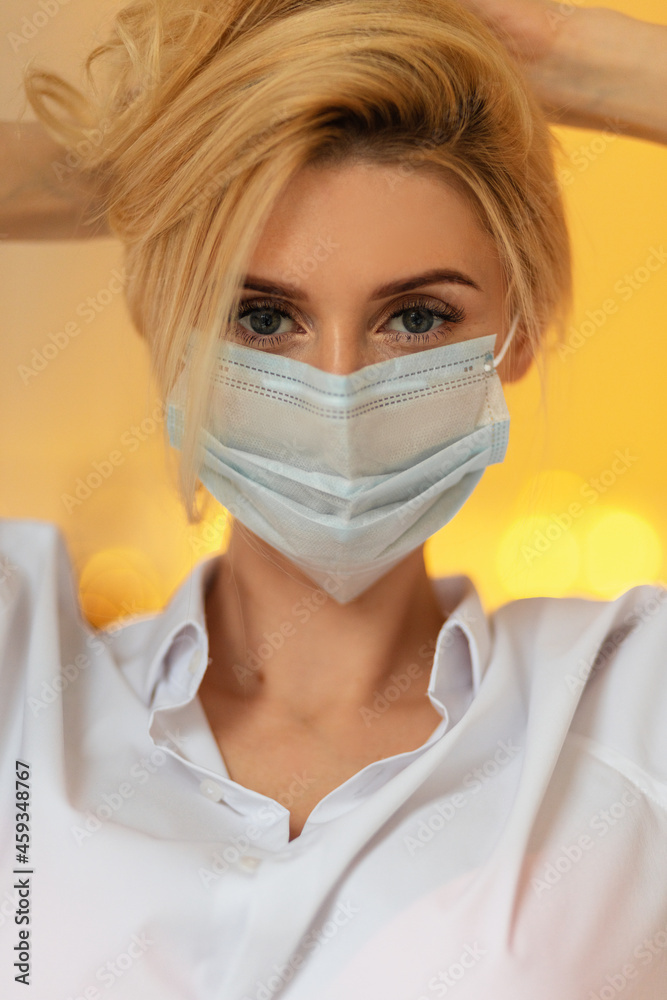 Close-up portrait of pretty young blonde woman with a medical mask in a stylish white shirt against a background of golden bokeh lights.  girl nurse worker look at the camera indoors