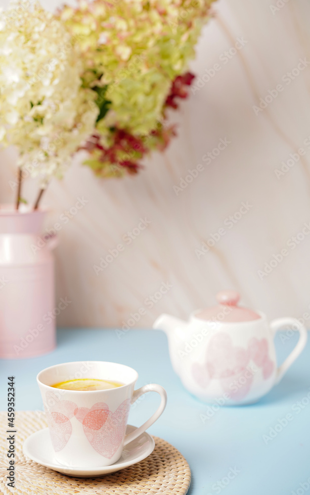 white cup with pink hearts a teapot and a bouquet of flowers on beige-blue background. Breakfast, morning tea. white cup with pink hearts a teapot and a bouquet of flowers on beige-blue background. Breakfast, morning tea.
