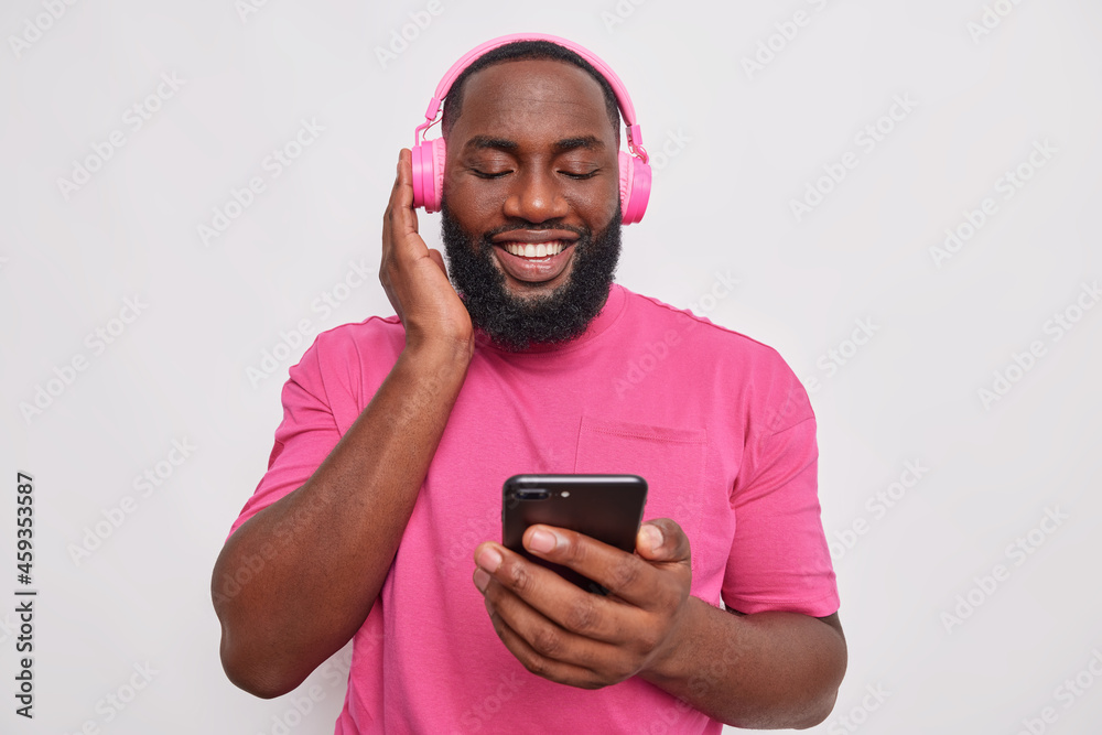 Positive dark skinned bearded man checks sound on his new headphones holds mobile phone chooses song to listen wears casual pink t shirt isolated over white background downloads application. Positive dark skinned bearded man checks sound on his new headphones holds mobile phone chooses song to listen wears casual pink t shirt isolated over white background downloads application.