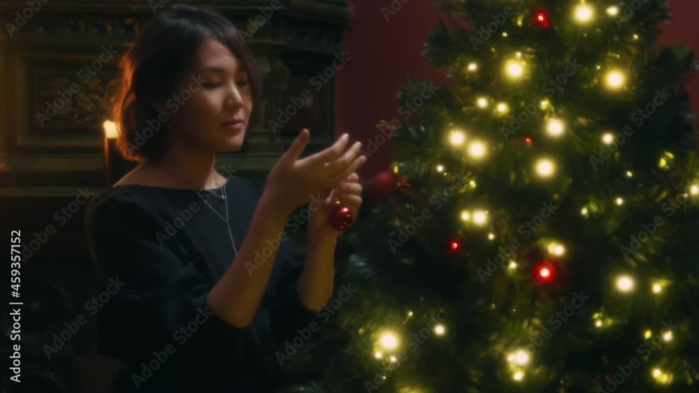 Young asian woman decorating Christmas tree with red sparkling ball. Attractive girl preparing to celebrate New Year at home, hanging toys to Christmas tree, illuminated by garlands and candles Young asian woman decorating Christmas tree with red sparkling ball. Attractive girl preparing to celebrate New Year at home, hanging toys to Christmas tree, illuminated by garlands and candles