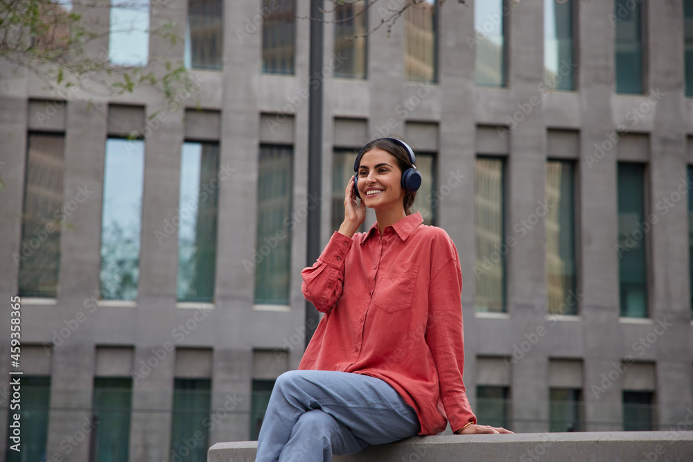 Fototapeta premium Cheerful beautiful woman sits relaxed against urban setting listens music in wireless headphones dressed in casual shirt and jeans enjoys music playlist popular audio tracks during leisure time.