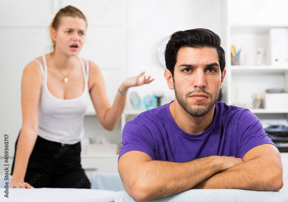 Fototapeta premium Frustrated guy sitting at home table on background with dissatisfied girlfriend