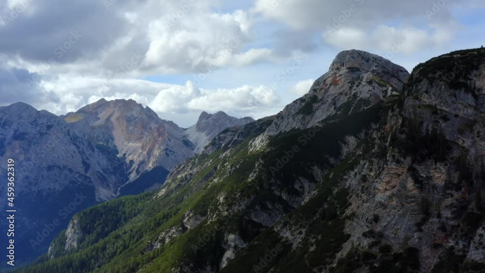 Dolomites, Cortina d'Ampezzo, Veneto, Italy, September 2021. Drone pushes towards densely wooded cliffs, with a vista of rugged mountains touching the clouds in the distance. Dolomites, Cortina d'Ampezzo, Veneto, Italy, September 2021. Drone pushes towards densely wooded cliffs, with a vista of rugged mountains touching the clouds in the distance.