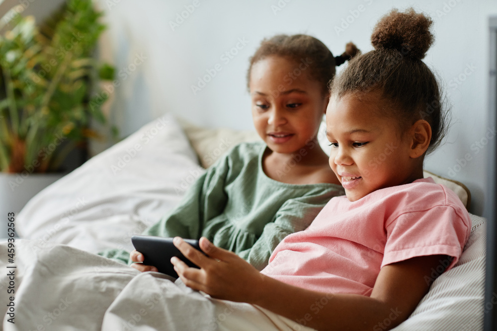 Portrait of two cute girls using smartphone together while sitting lying on bed in cozy room and watching videos Portrait of two cute girls using smartphone together while sitting lying on bed in cozy room and watching videos