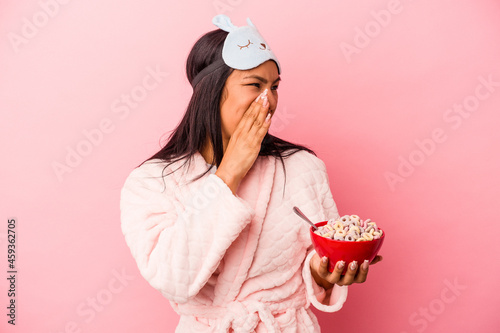 Young latin woman wearing pajama holding a bowl of cereals isolated on pink background  shouting and holding palm near opened mouth.