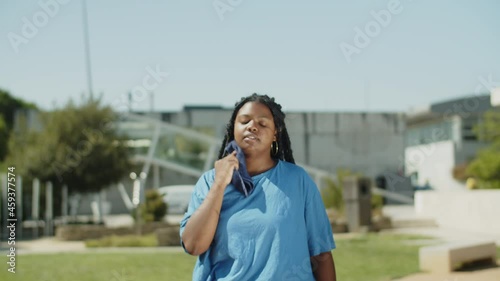 Tired woman wiping sweat with towel after training in park. Tracking shot of African-American fat person finished cardio training at athlete stadium and drinking water. Body positive, sport concept photo