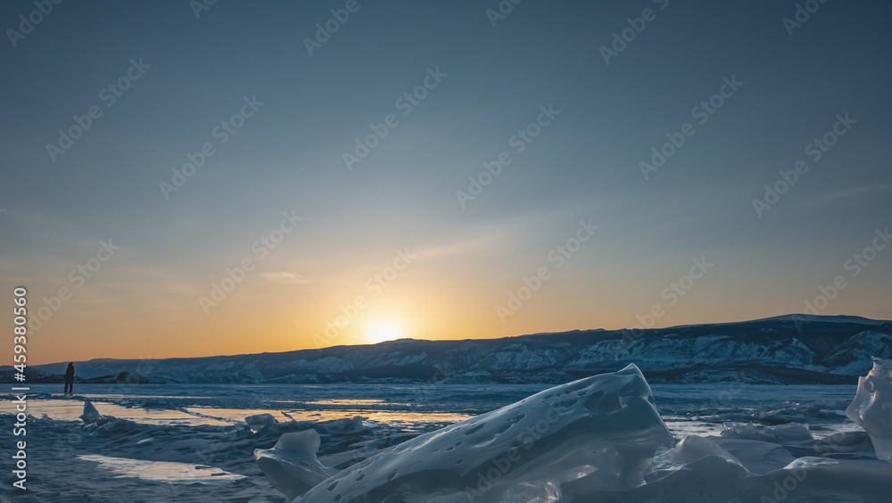 Winter evening. The sun is hiding behind the mountains, the sky is highlighted orange. Glare on a frozen lake. The silhouette of a lone man on the ice. In the foreground - a block of hummocks. Baikal