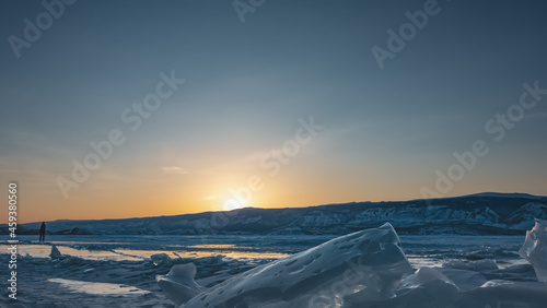 Winter evening. The sun is hiding behind the mountains, the sky is highlighted orange. Glare on a frozen lake. The silhouette of a lone man on the ice. In the foreground - a block of hummocks. Baikal © Вера 